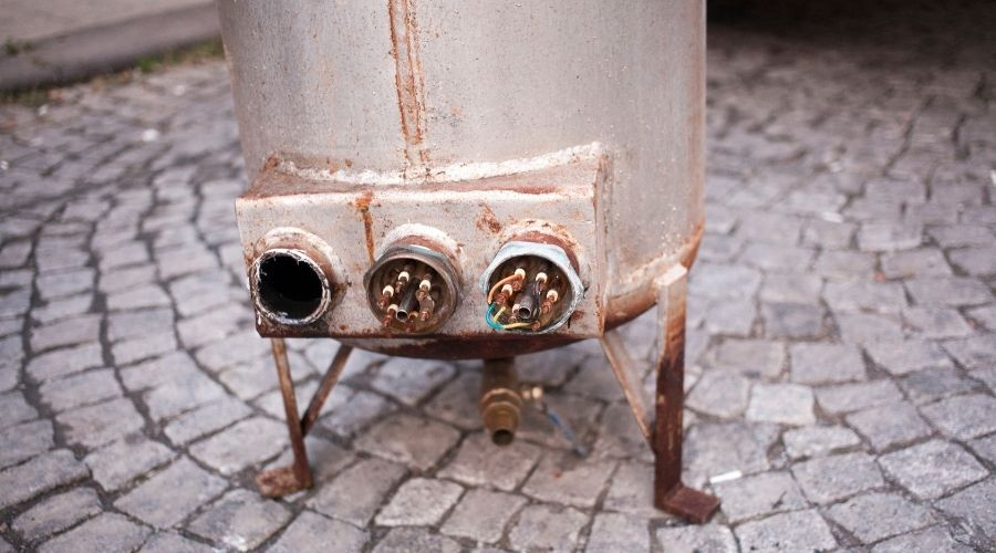Old, rusty water heater with visible pipes and connections, situated on cobblestone surface, relevant to plumbing maintenance and replacement discussions.