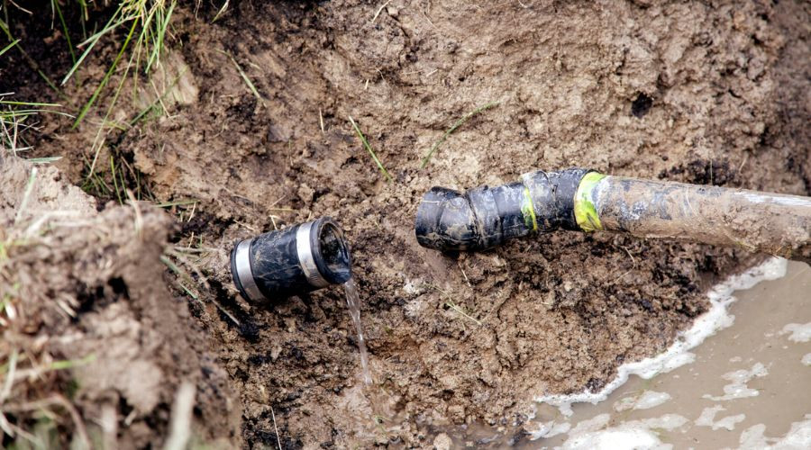 Pipe leaking water from a sewer line in muddy ground, indicating potential plumbing issues and the need for repair or replacement.